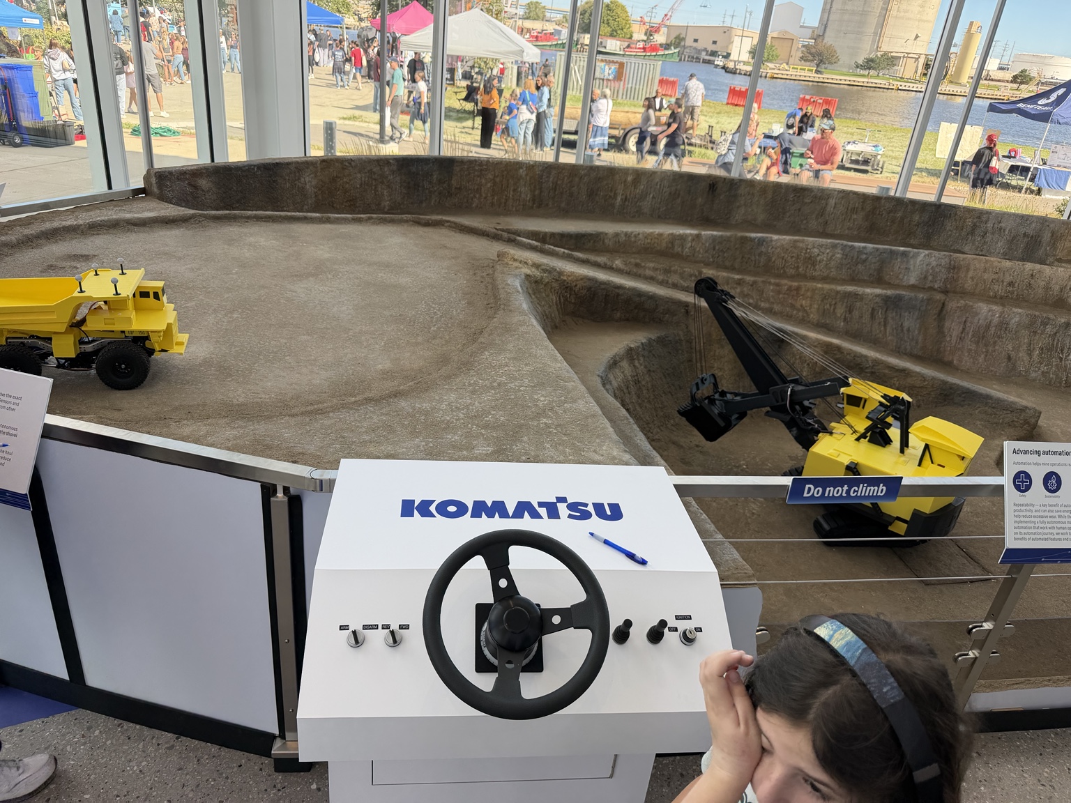 A Komatsu steering-wheel control console overlooking a model mine with yellow haul trucks and a mining shovel.